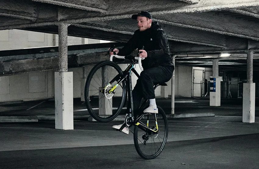 man wearing black clothes doing a wheelie on a road bike in a parking garage